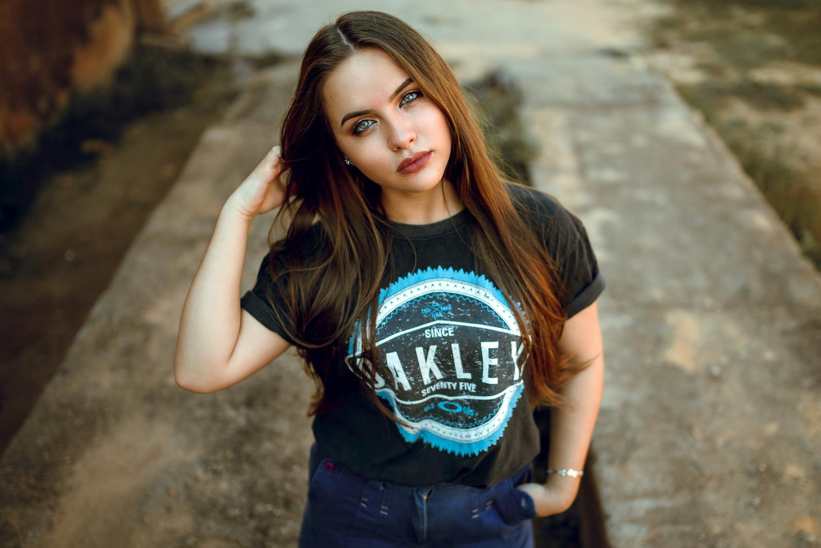 A young woman with long brunette hair poses confidently outdoors in casual attire.